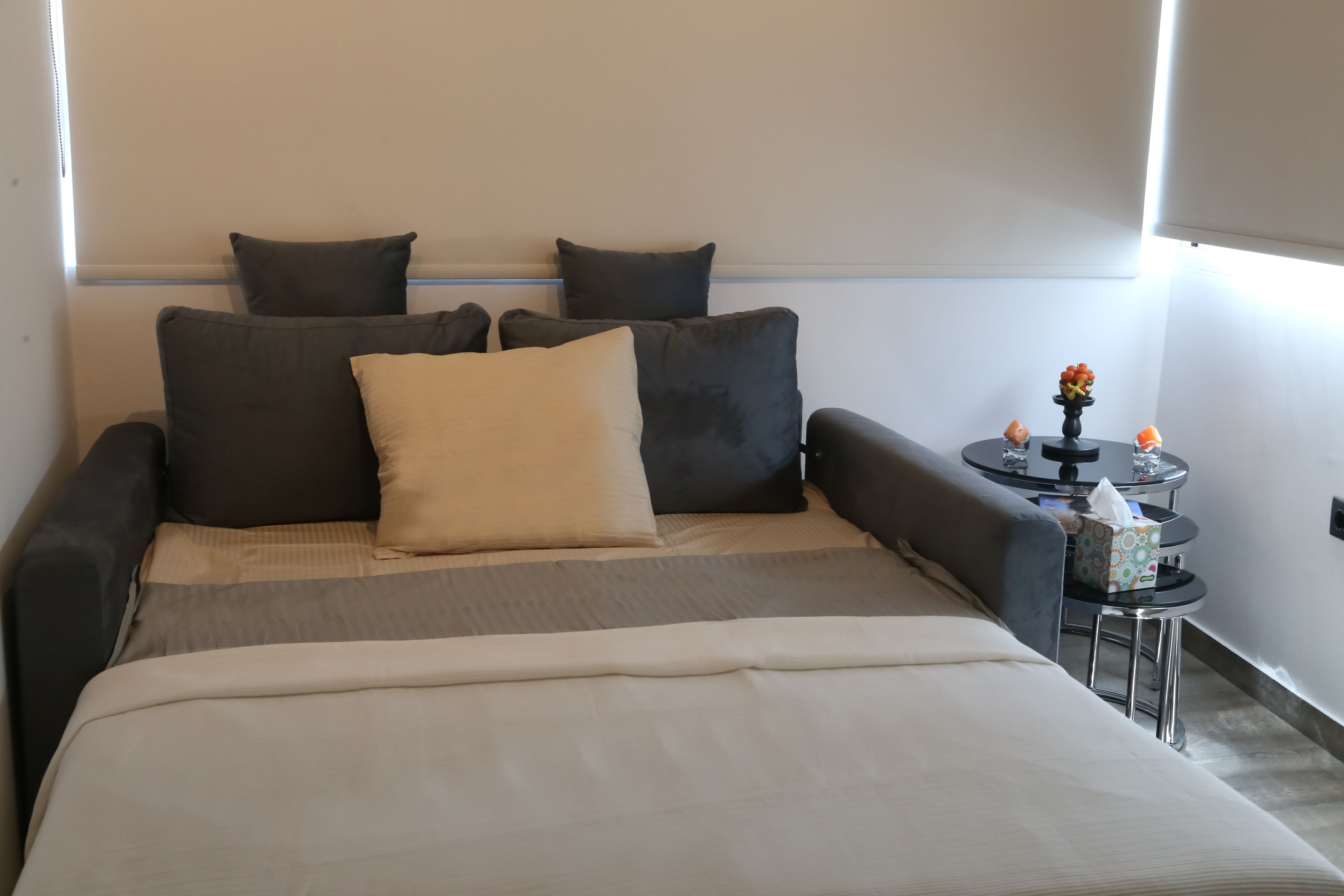Un lit bien fait, recouvert d’une couette beige et agrémenté de plusieurs coussins, une petite table ronde sur laquelle est posé un vase de fleurs, et une boîte de mouchoirs.