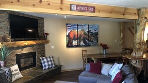 Un salon chaleureux avec une cheminée en pierre, un téléviseur à écran plat fixé au-dessus, et un coin repas avec une table et des chaises en bois.