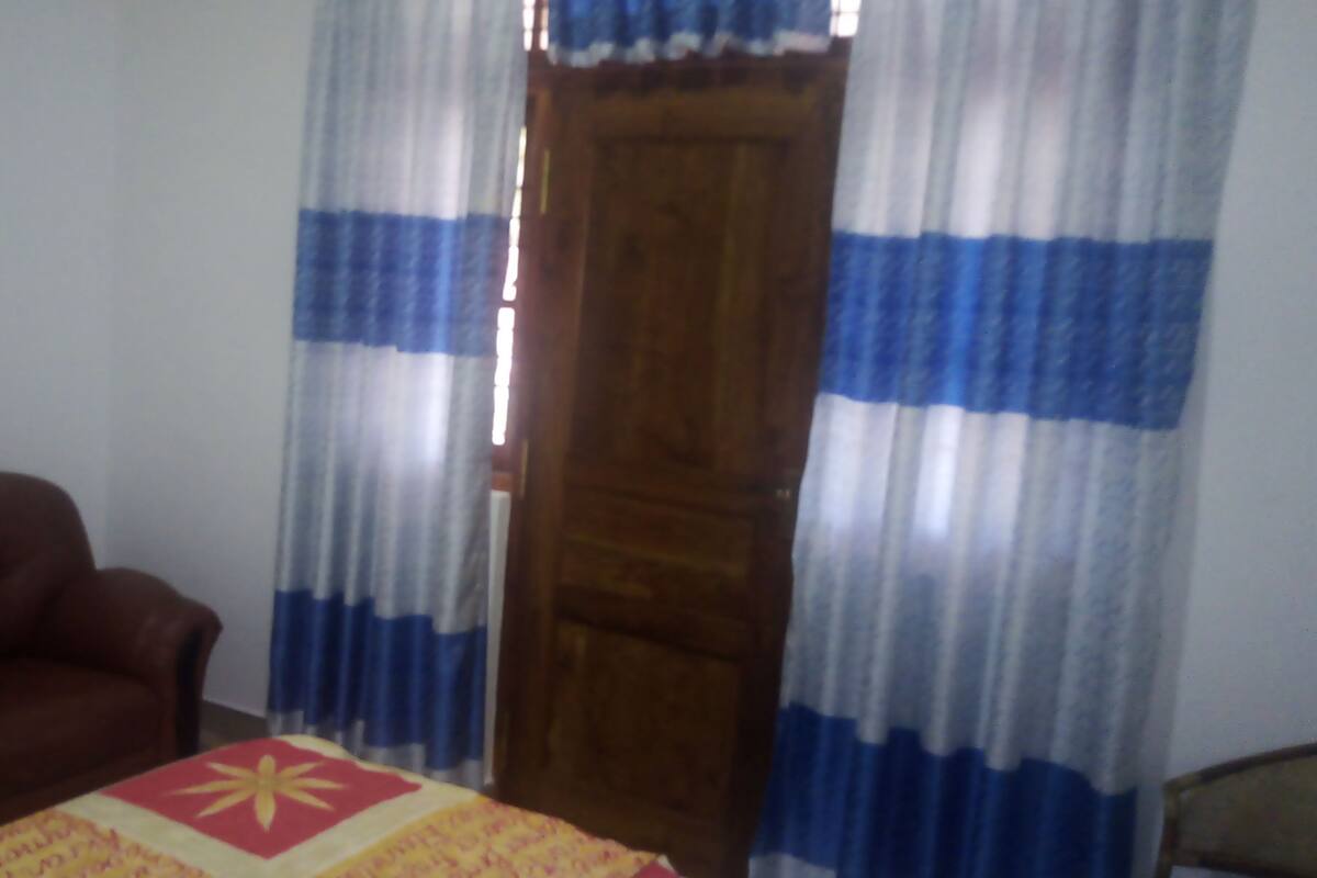 Ein Zimmer mit einem braunen Ledersessel, einem Bett mit einer roten und gelben Steudecke und blau-weißen, gestreiften Vorhängen.