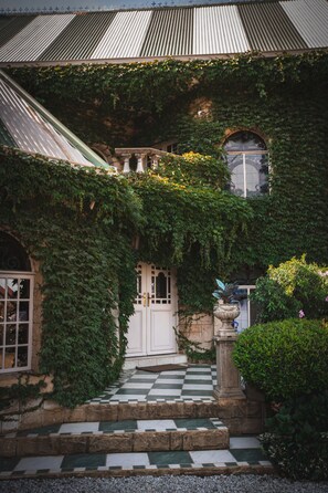 A building with a checkered stone pathway leading to a white door, surrounded by lush greenery and ivy. - Schoon Huis Manor (Kempton Park)