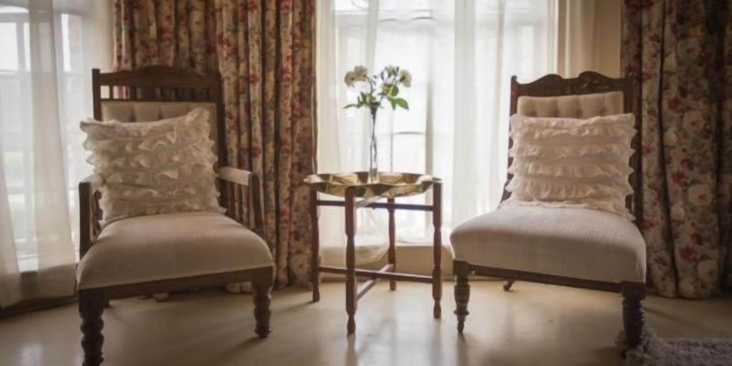 Two upholstered chairs with ruffled pillows, a small round table with a vase, and curtains with a floral pattern.