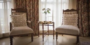 Two upholstered chairs with ruffled pillows, a small round table with a vase, and curtains with a floral pattern. - Schoon Huis Manor (Kempton Park)