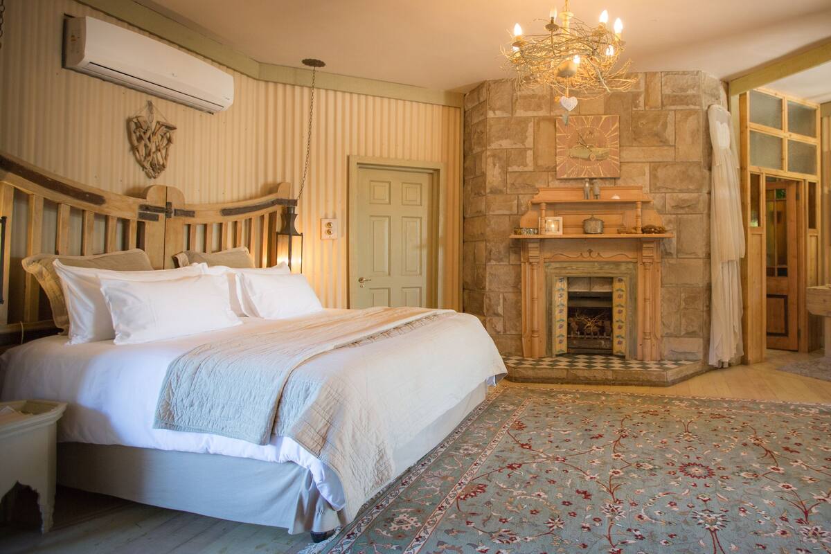 Une chambre avec un grand lit, une cheminée en pierre, une commode en bois et un miroir.