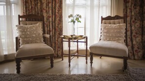 Deux fauteuils rembourrés ornés de coussins à volants, une petite table ronde sur laquelle est posé un vase de fleurs, et des rideaux à motifs floraux.