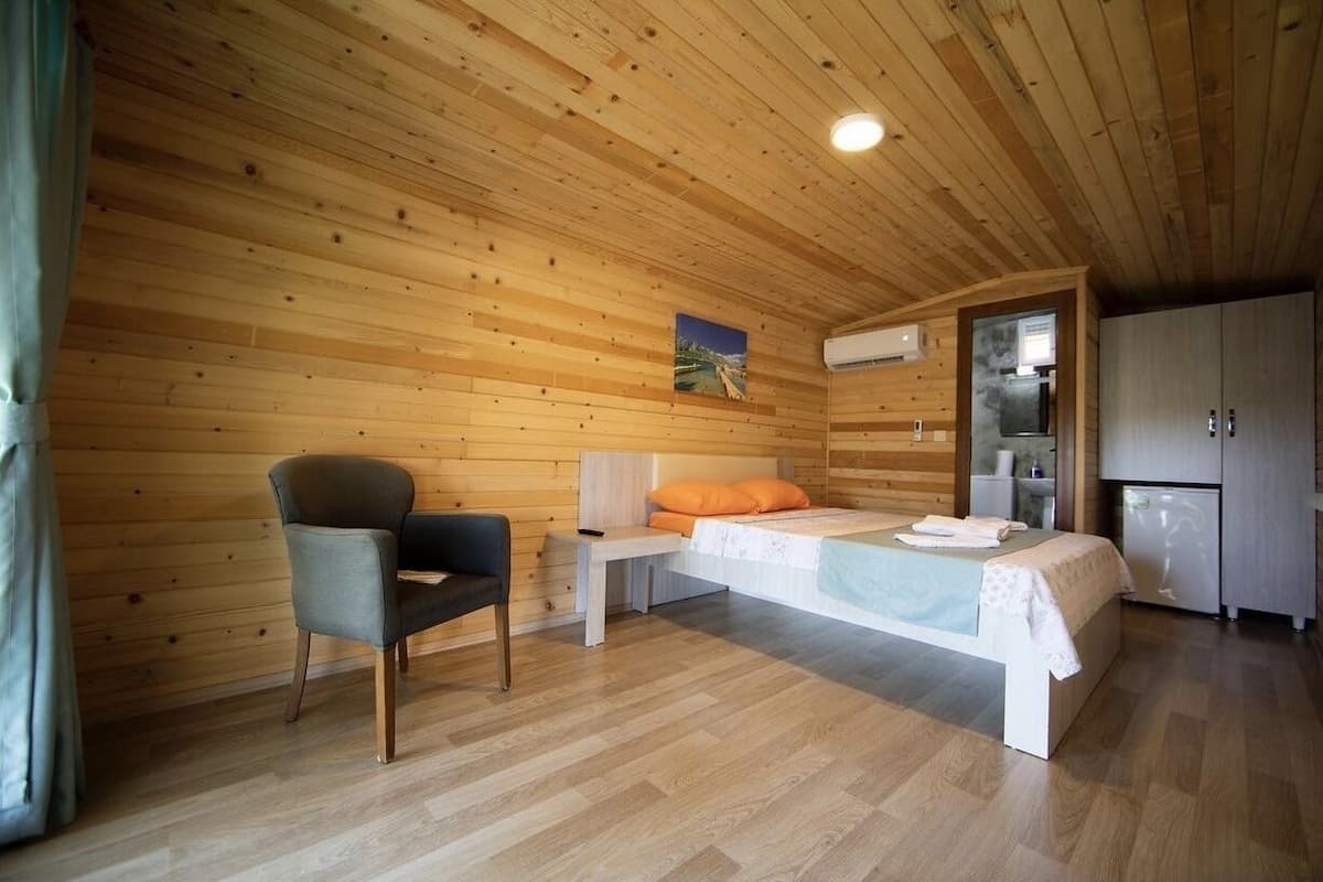 A wooden cabin room with a bed, a chair, a small table, and a kitchenette.