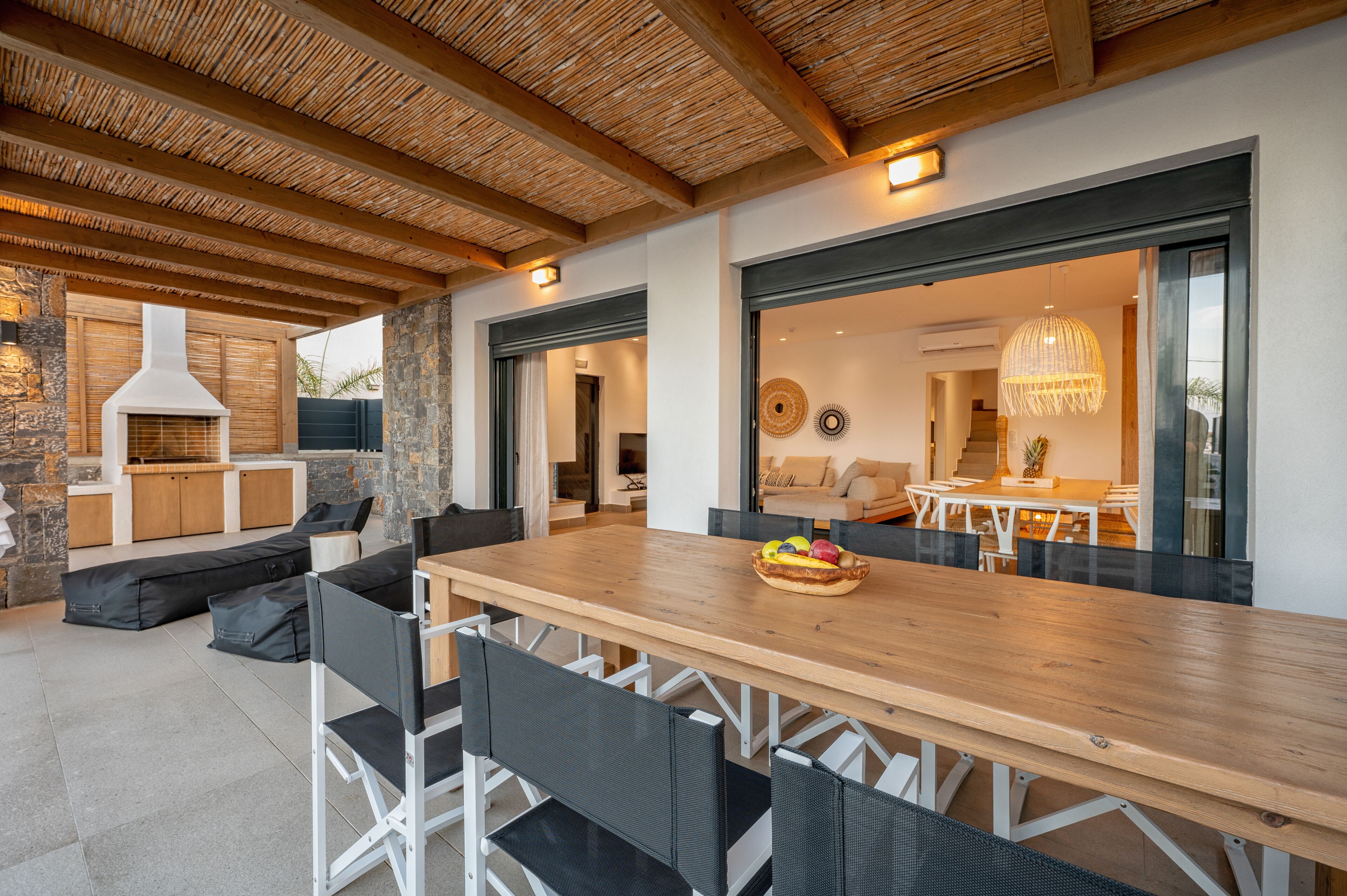 An outdoor dining area with a wooden table and chairs, a stone fireplace, and a view into the house.