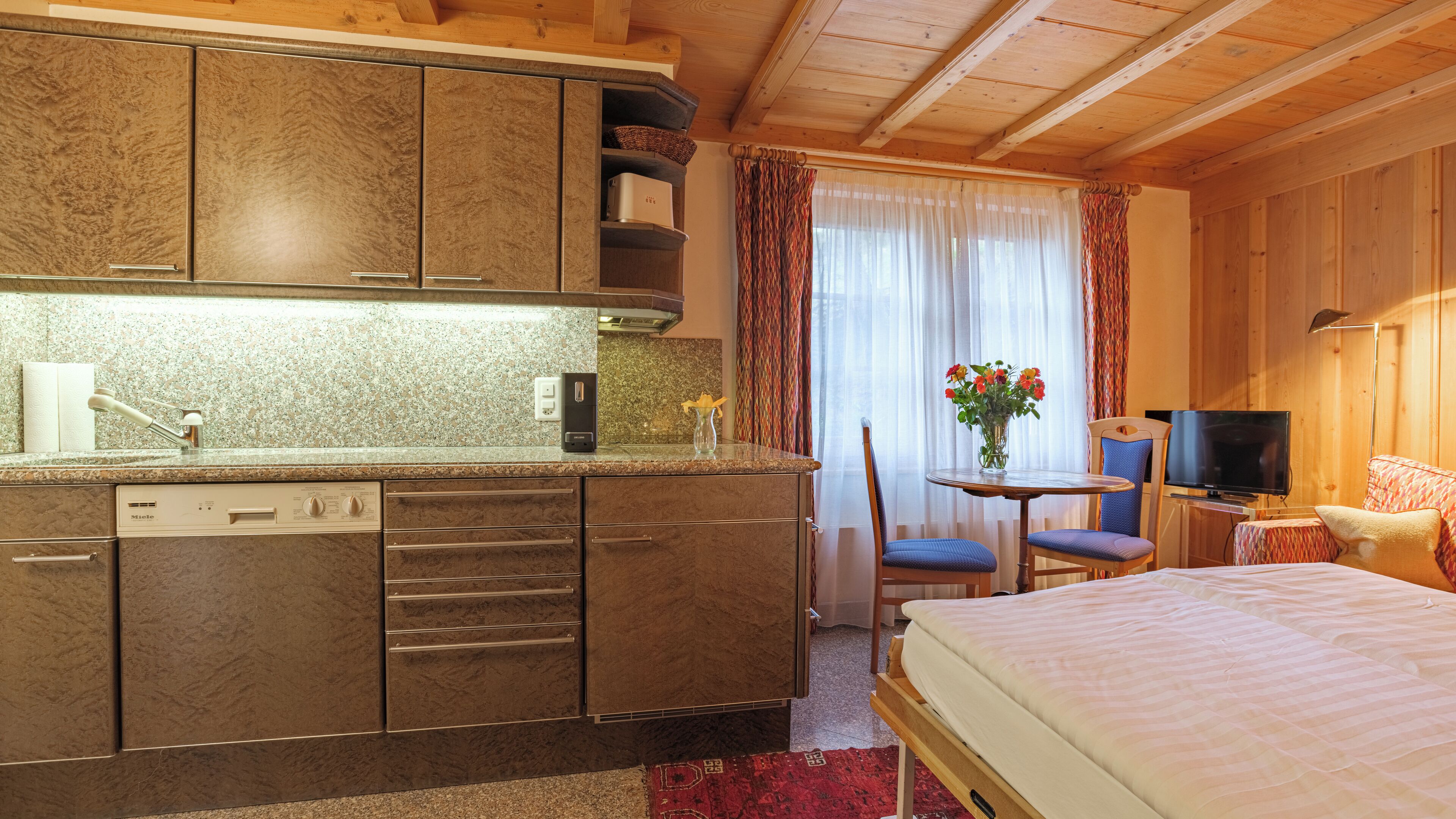 A compact hotel room with a kitchenette, a bed, a TV, and a small dining area.