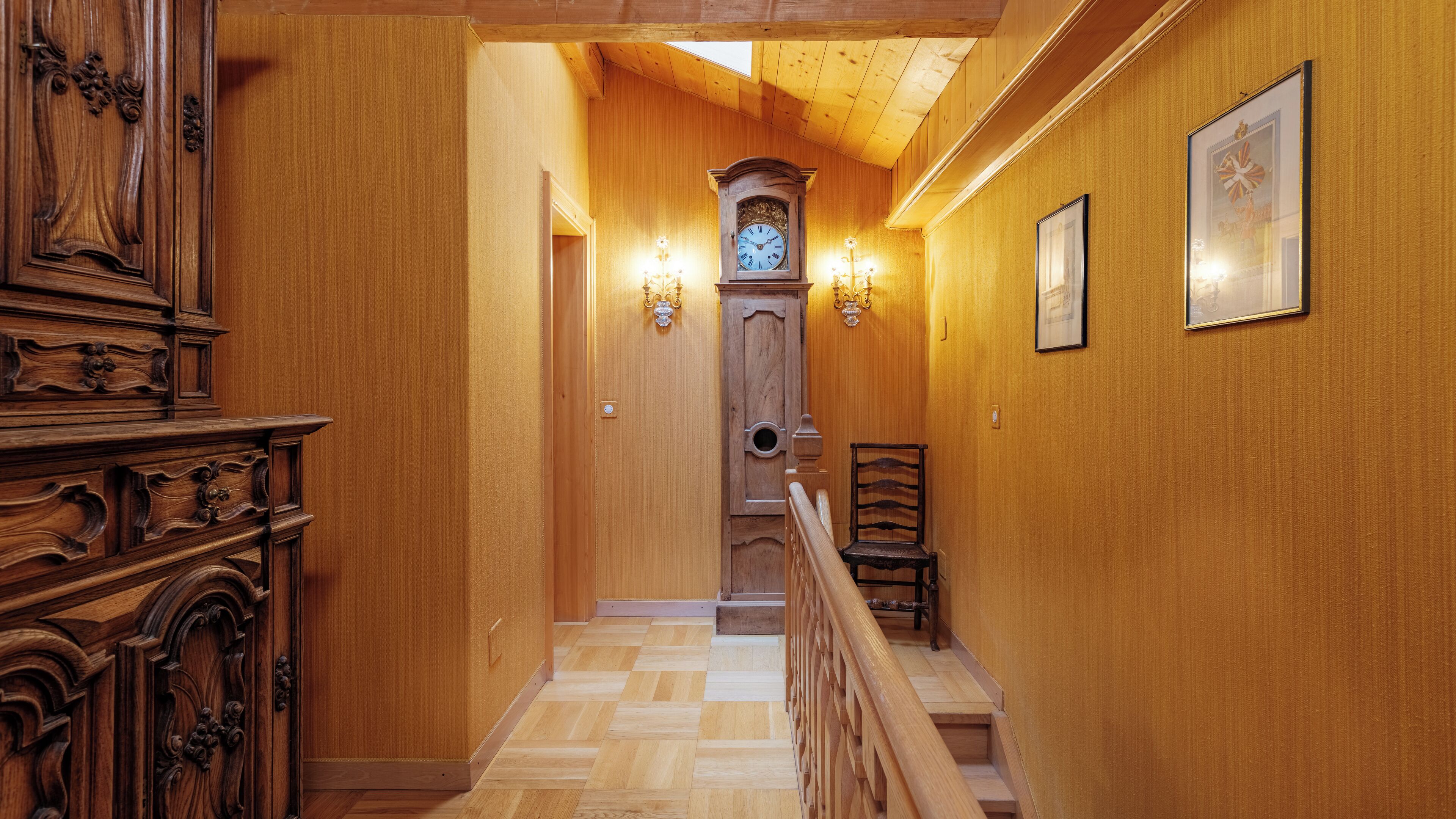 A wooden hallway with a wooden staircase, a wall-mounted clock, and two wall sconces.