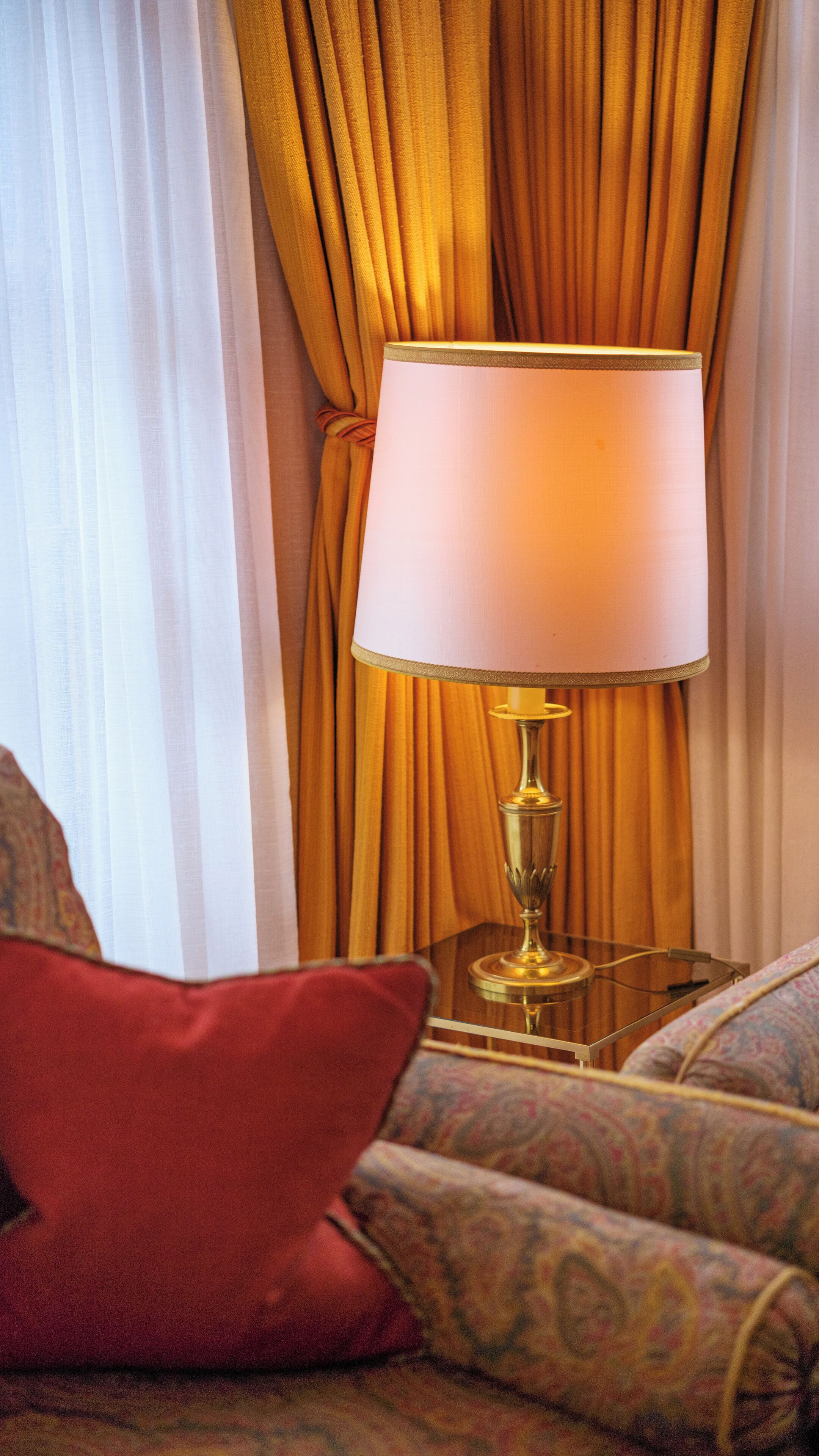 A room with a lamp on a glass table, a red pillow, and patterned upholstery.