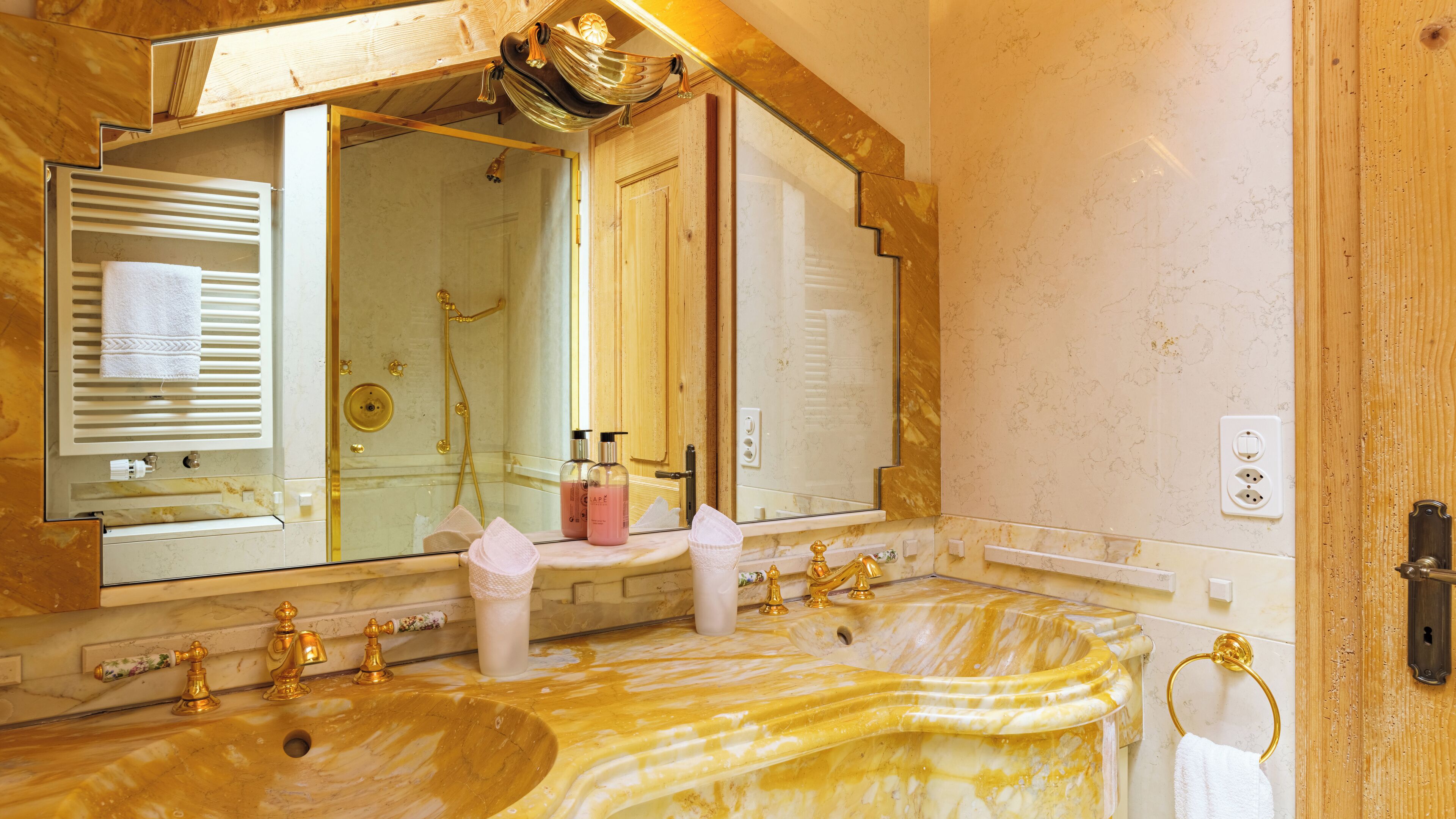 Une salle de bain dotée d’un grand miroir, de deux lavabos avec des robinets dorés, d’une douche et d’un porte-serviettes.