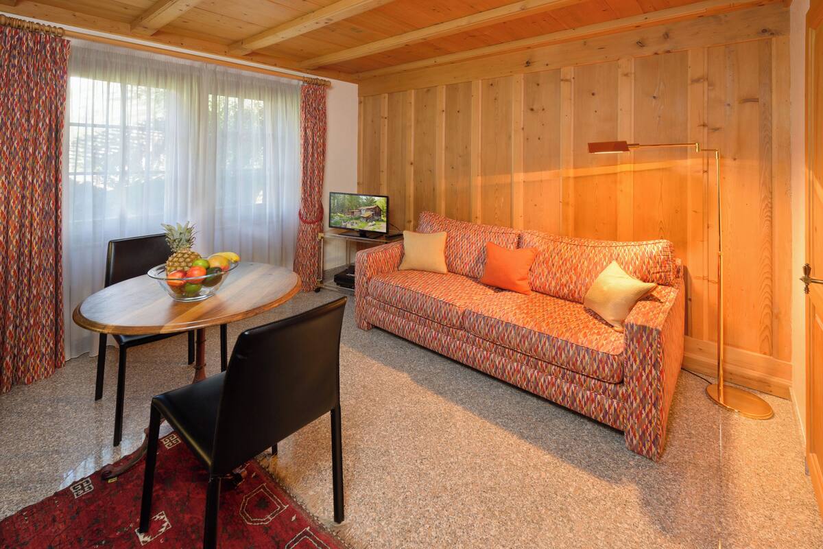 A cozy living room with a wooden ceiling, a patterned sofa, a dining table with chairs, and a television.