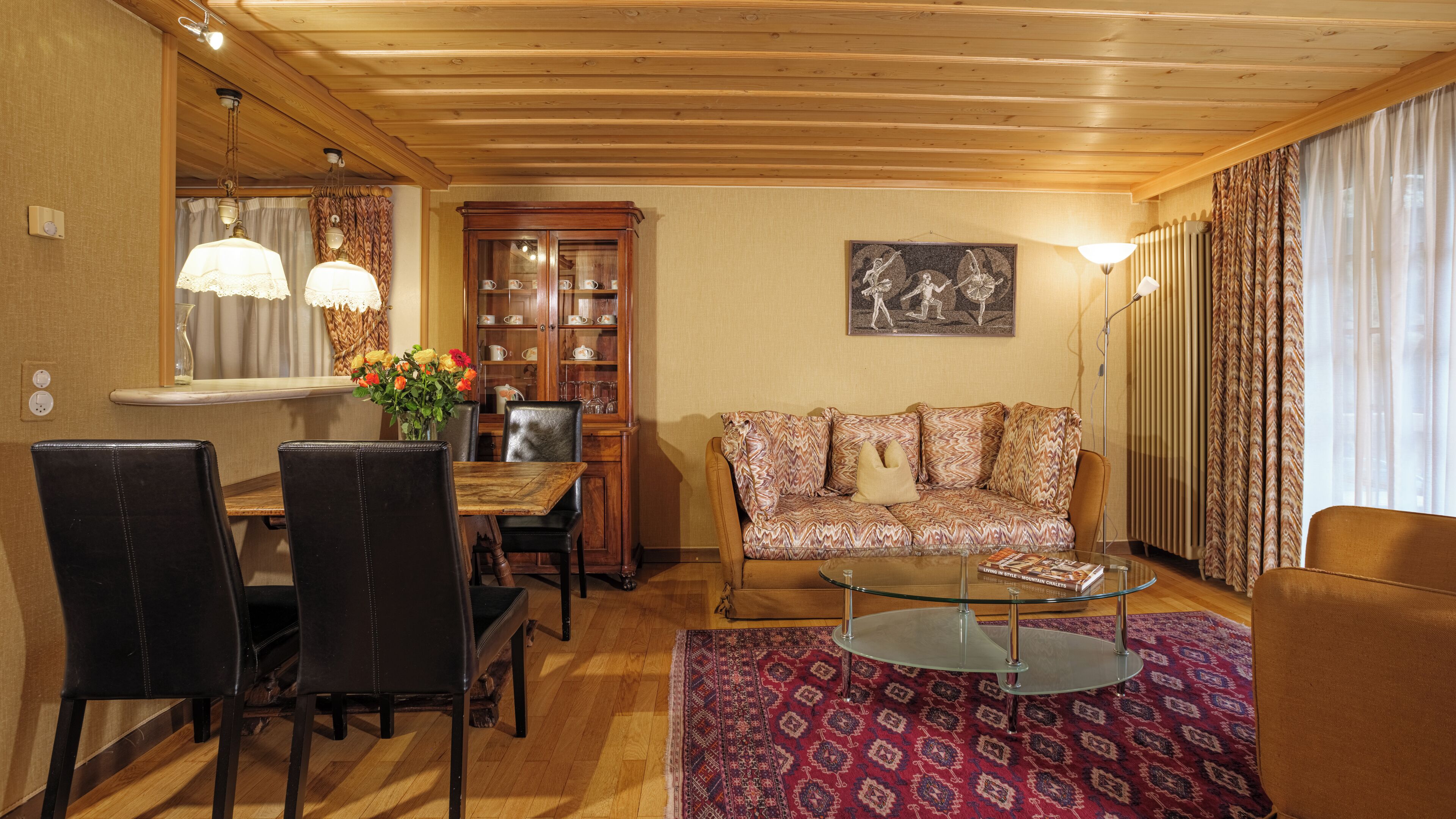 Uma sala de estar aconchegante com teto de madeira, sofá estampado, mesa de centro de vidro e um espaço de jantar com cadeiras e um armário.