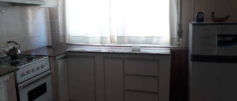 Una cocina con armarios blancos, estufa, refrigerador y una ventana con cortinas ligeras.