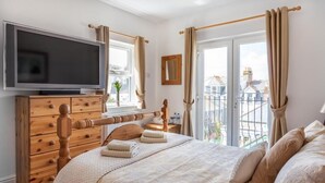 Une chambre d’hôtel avec une commode en bois, un téléviseur à écran plat, un lit avec du linge de lit beige et un balcon avec des rideaux.