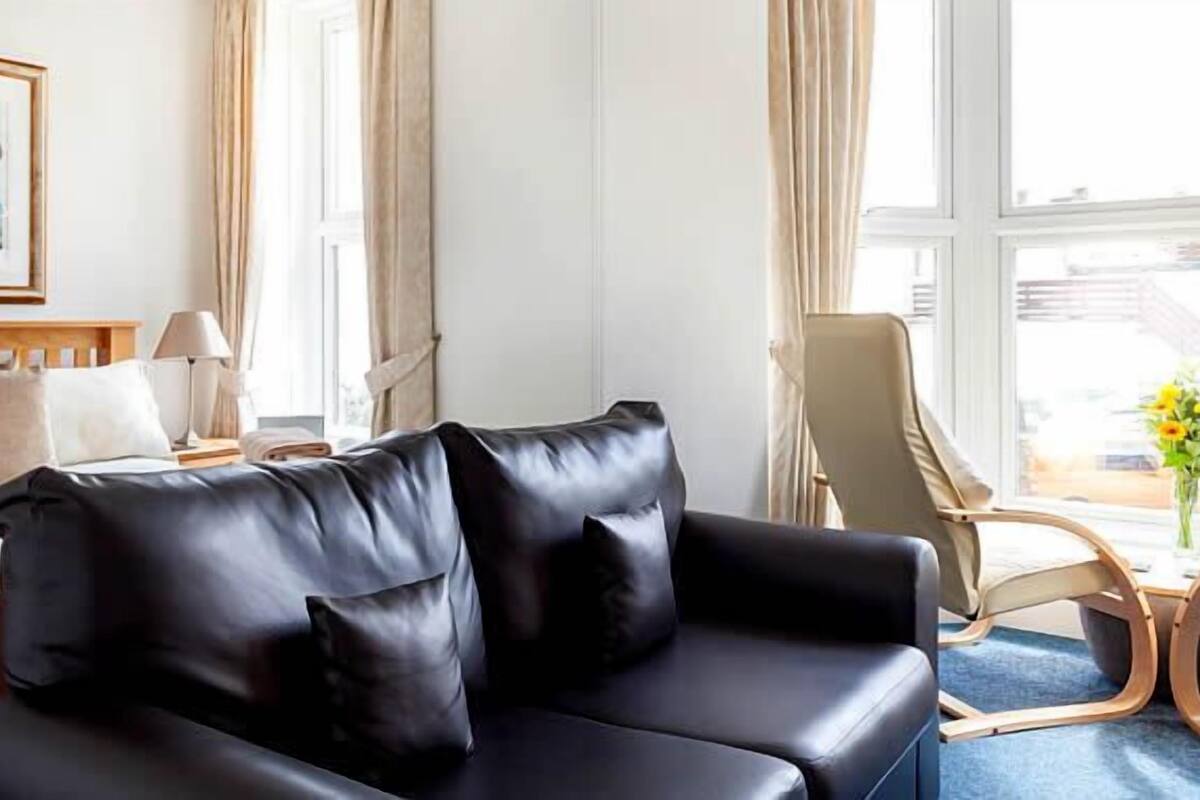 A hotel room with a bed, a black leather sofa, a wooden nightstand, a lamp, and a window with a view of the street.