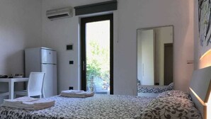 Habitació d'hotel amb llit, escriptori, cadira, nevera i finestra amb vista a vegetació.