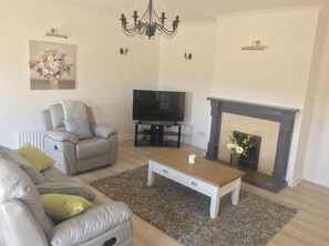 Ein Wohnzimmer mit grauem Sofa, Couchtisch, Fernseher, Kamin und einer Chandelier.