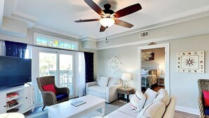 A living room with a ceiling fan, a TV, a sofa, a coffee table, and a wicker chair. - Bay View Villas 105 | Waterfront, Community Pool (Tybee Island)