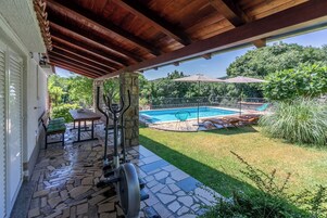 A covered patio with a stone floor, outdoor exercise equipment, a pool, and a view of a lush garden.