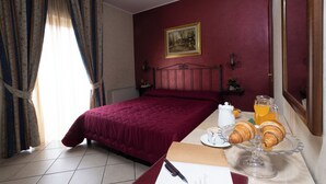 A hotel room with a bed, a bedside table with a coffee cup and croissants, a painting on the wall, and curtains. - Hotel Gangi (Piazza Armerina)