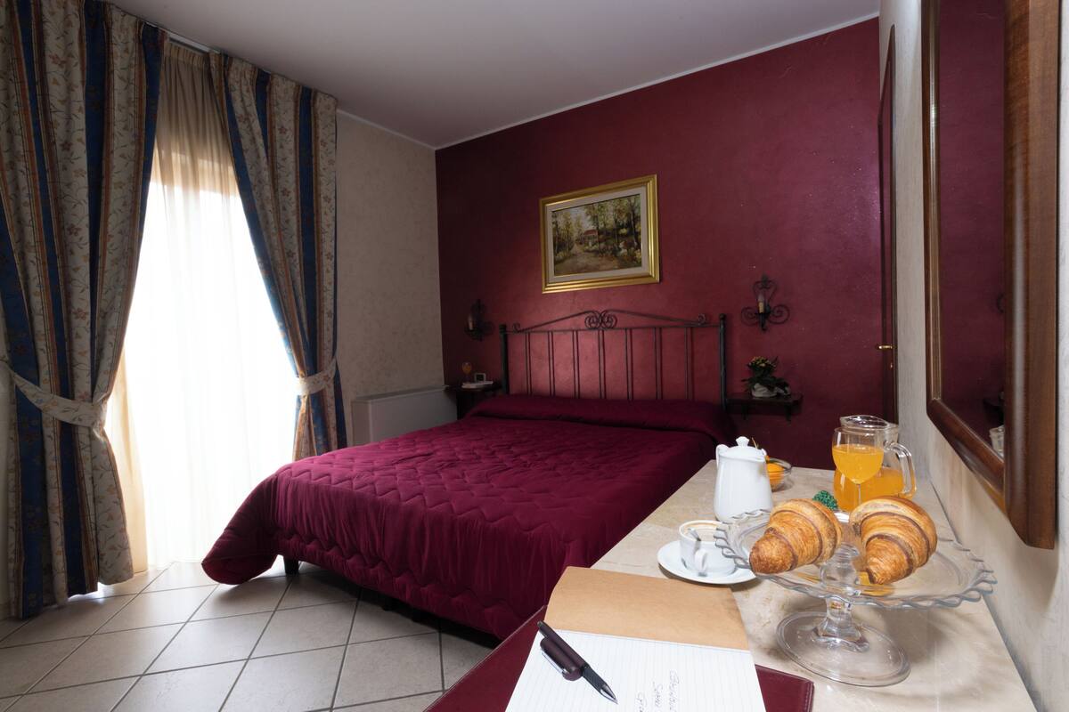 A hotel room with a bed, a bedside table with a coffee cup and croissants, a painting on the wall, and curtains.