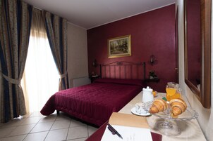 Una habitación de hotel con una cama, una mesita de noche con una taza de café y croissants, un cuadro en la pared y cortinas.