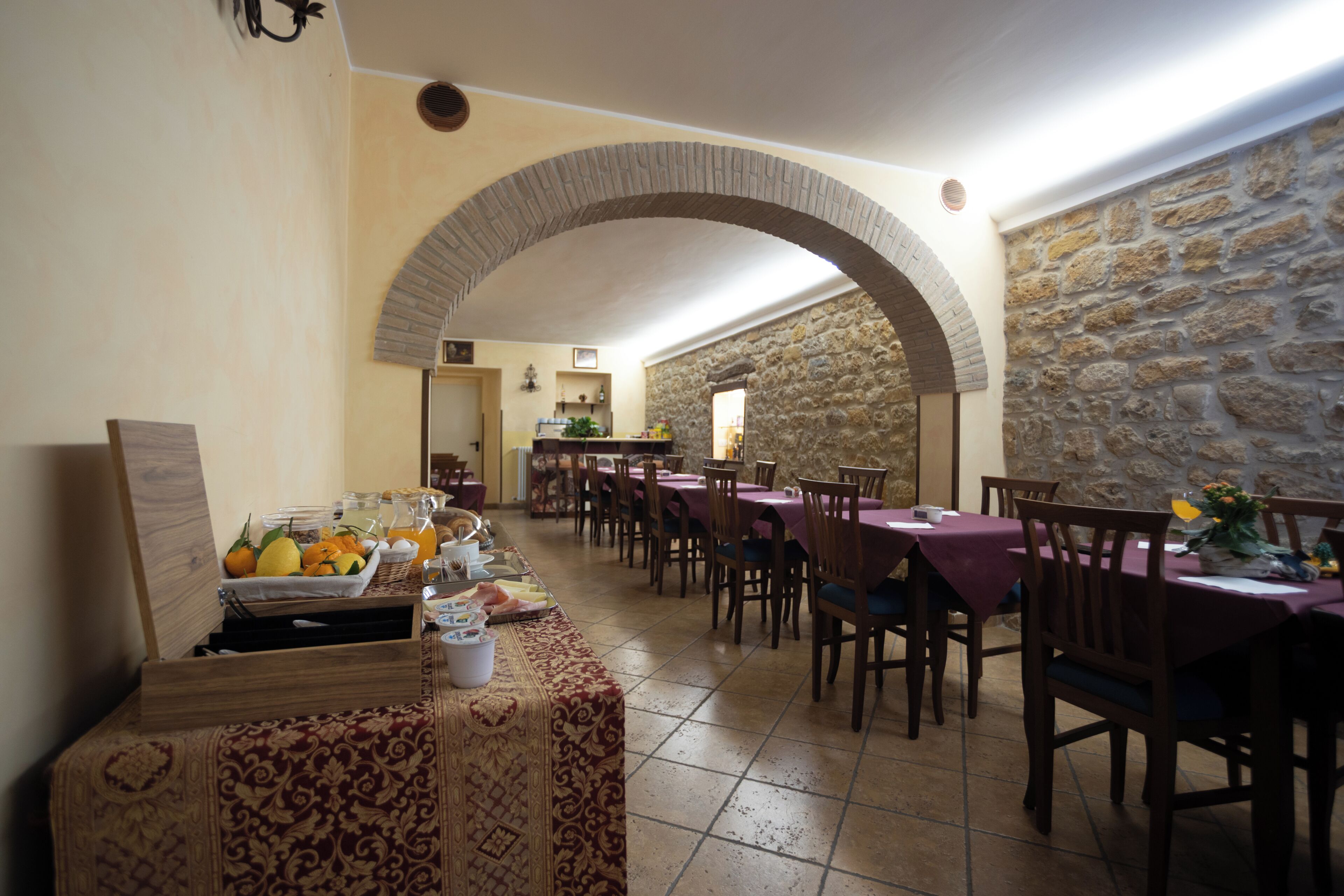 A dining area with tables and chairs, a buffet with fruits and drinks, and a stone wall.