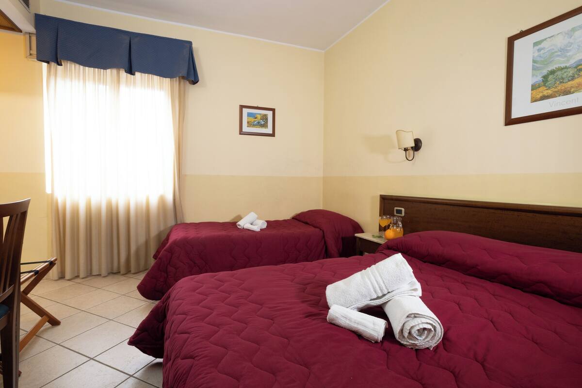 A hotel room with two beds, a chair, a desk, a window with curtains, a wall-mounted picture, and a nightstand with a lamp and a bottle.