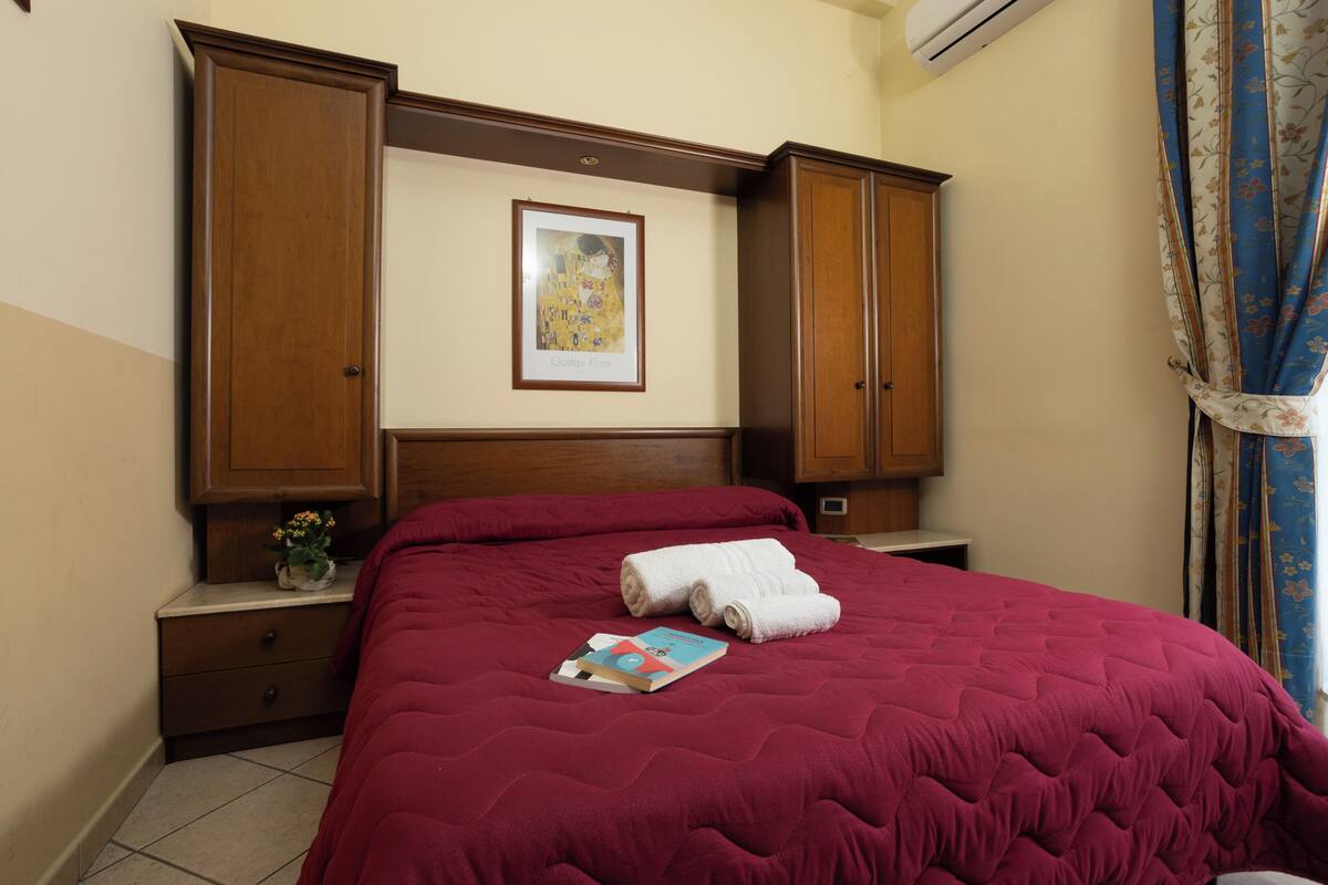A hotel room with a large bed, a wooden headboard, a framed artwork, a nightstand, and a window with curtains.