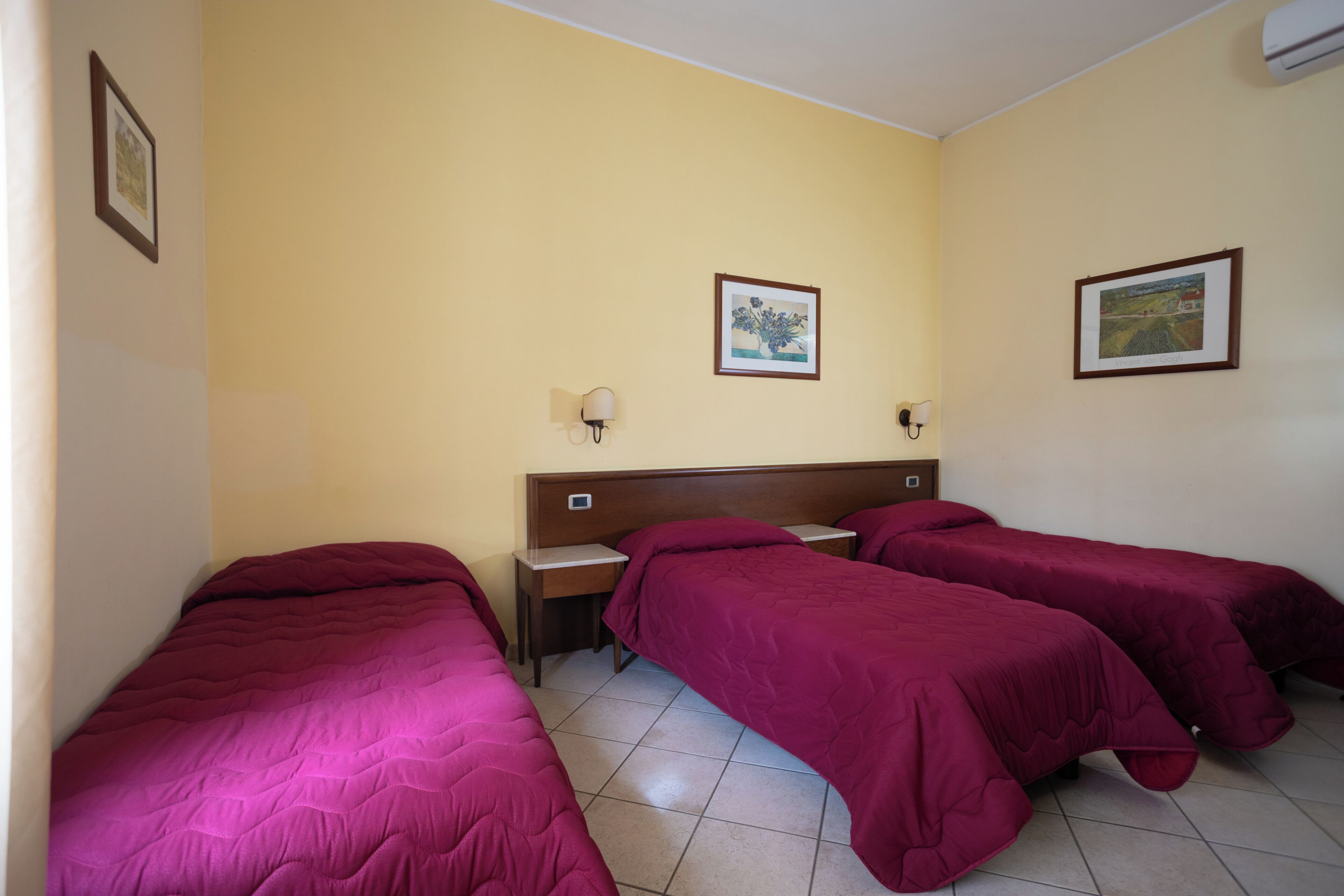 A hotel room with three single beds, a wooden headboard, two bedside tables, and two wall-mounted lamps.