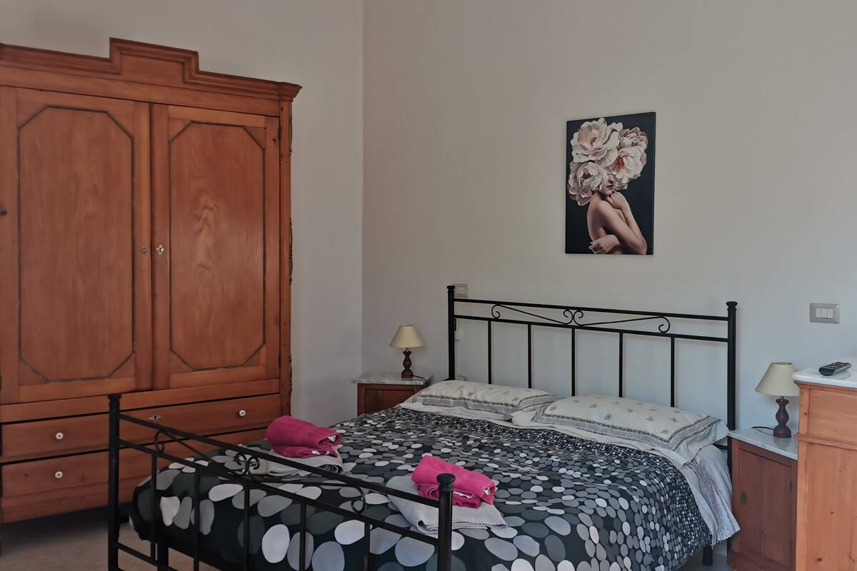 A bedroom with a bed, a wooden wardrobe, a nightstand, and a wall-mounted lamp.