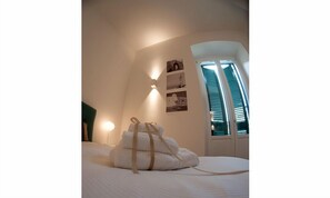 Una cama bien hecha con un juego de toallas, una lámpara de pared y una ventana con persianas.