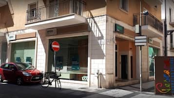 Un'auto rossa parcheggiata sulla strada di fronte a un edificio con un cartello 'BPER', un segnale di divieto di accesso e un cartello 'Gecara Shopping Village'.