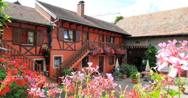A traditional half-timbered building with a staircase leading to a balcony, surrounded by vibrant flowers and potted plants.