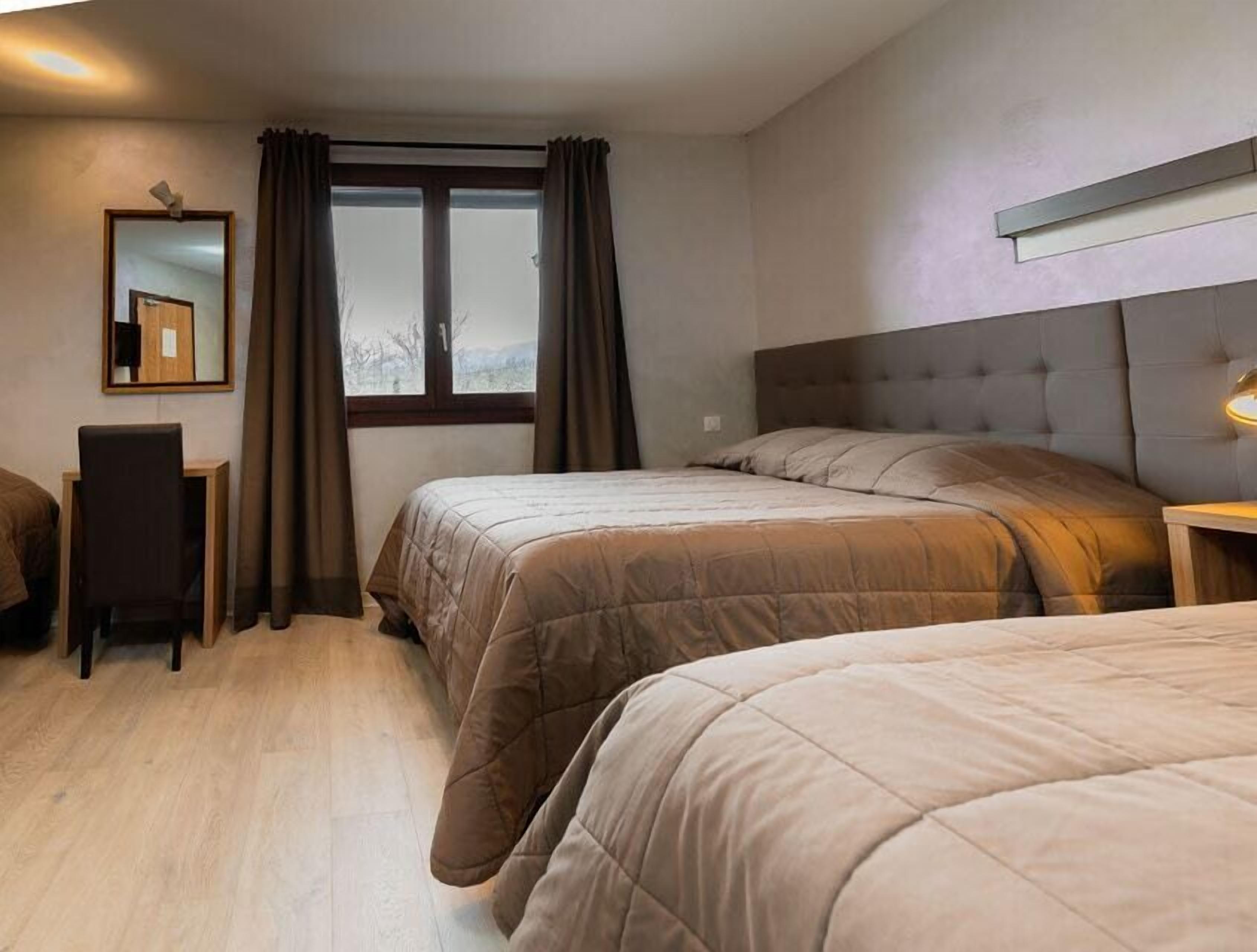 Une chambre d’hôtel avec deux lits, un bureau, une chaise, une fenêtre avec des rideaux et un miroir au mur.