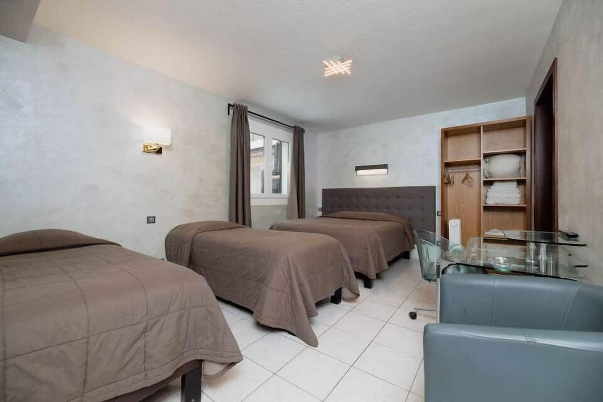 A hotel room with three single beds, a glass table, a grey armchair, and a wardrobe with shelves.