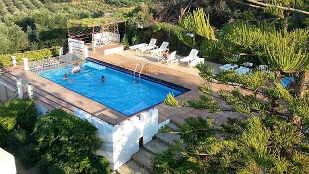 Uma piscina retangular com deck de madeira, rodeada por espreguiçadeiras brancas e vegetação.