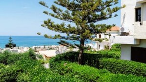 Um edifício moderno com fachada branca, um pinheiro em primeiro plano e vista para o mar e outros edifícios ao fundo.