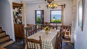 Uma sala de jantar com uma mesa posta para quatro pessoas, um candelabro e vista para o mar através de duas janelas.