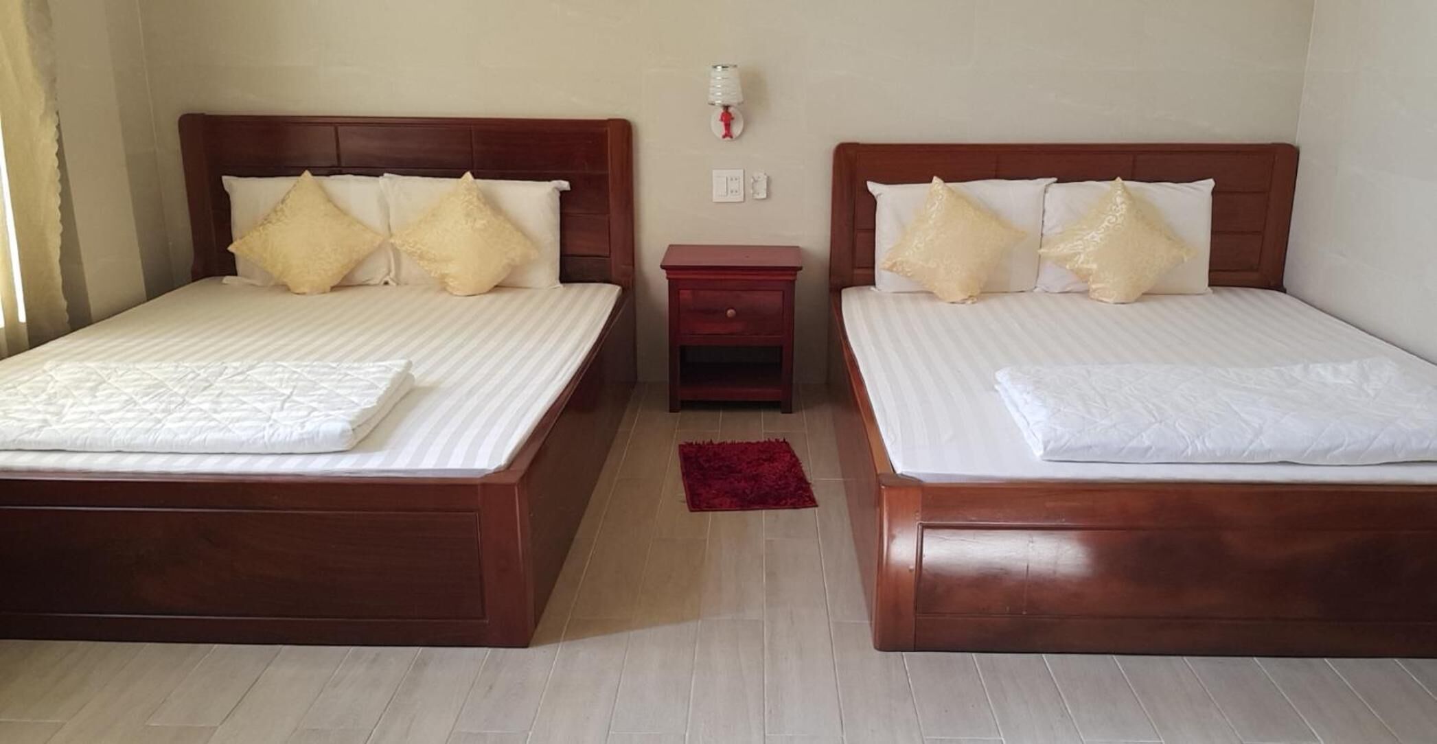 A hotel room with two beds, each with a wooden headboard, white bedding, and matching pillows. There is a small wooden nightstand between the beds, and a red rug on the floor.