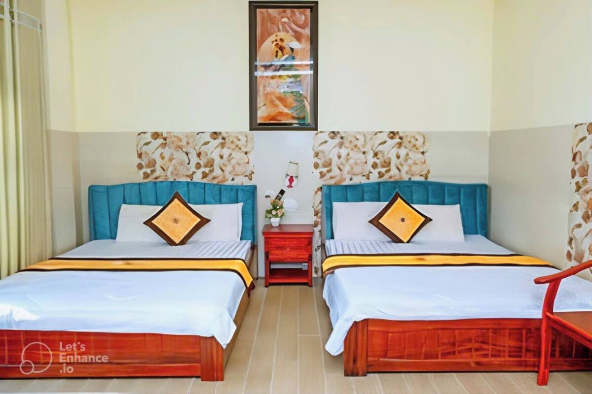 A hotel room with two beds, each with a blue headboard and a wooden frame. There is a small red nightstand between the beds with a vase of flowers.
