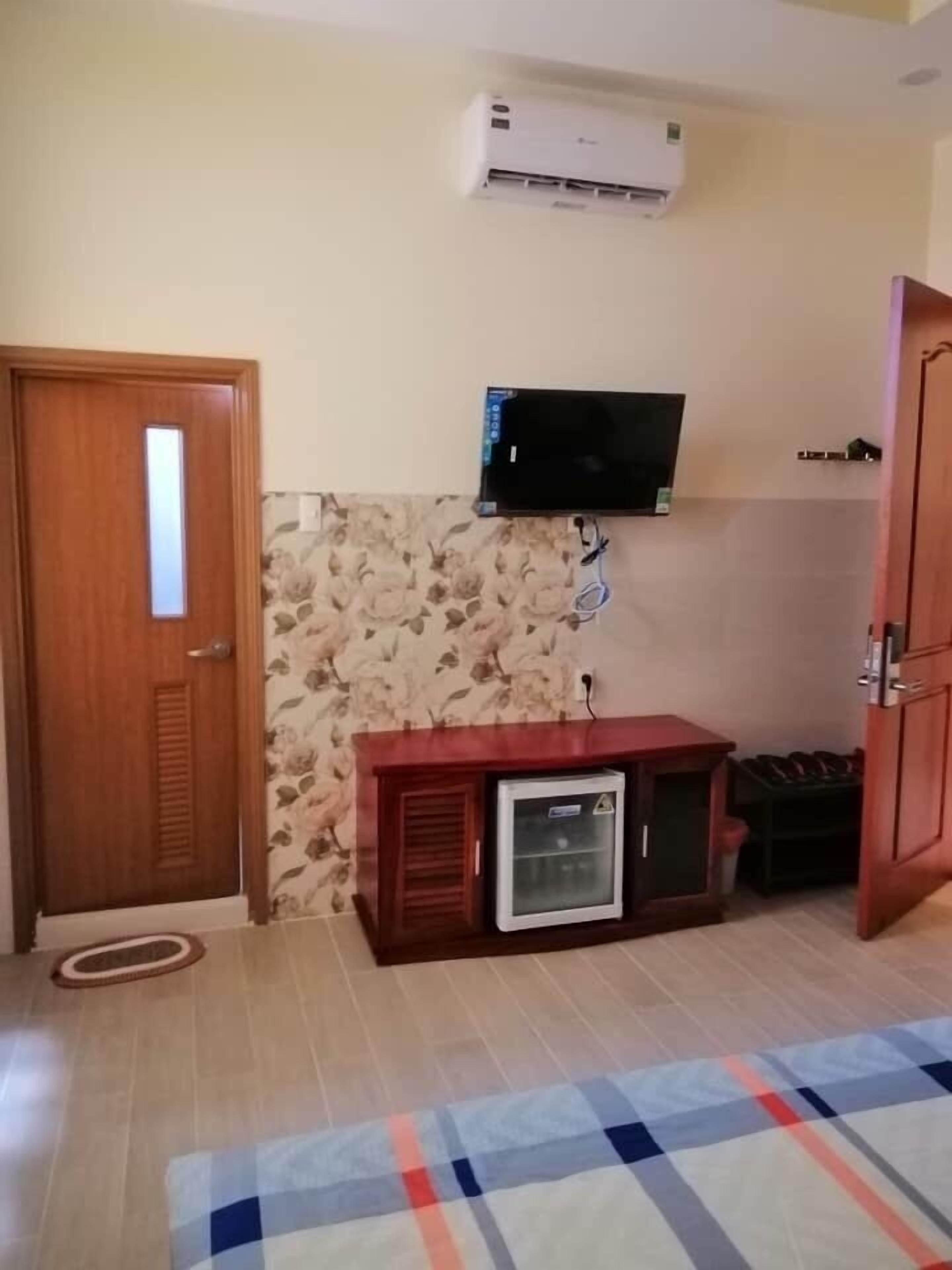Quarto com porta de madeira, pequeno frigorífico, televisão fixada na parede e tapete com padrão no chão.