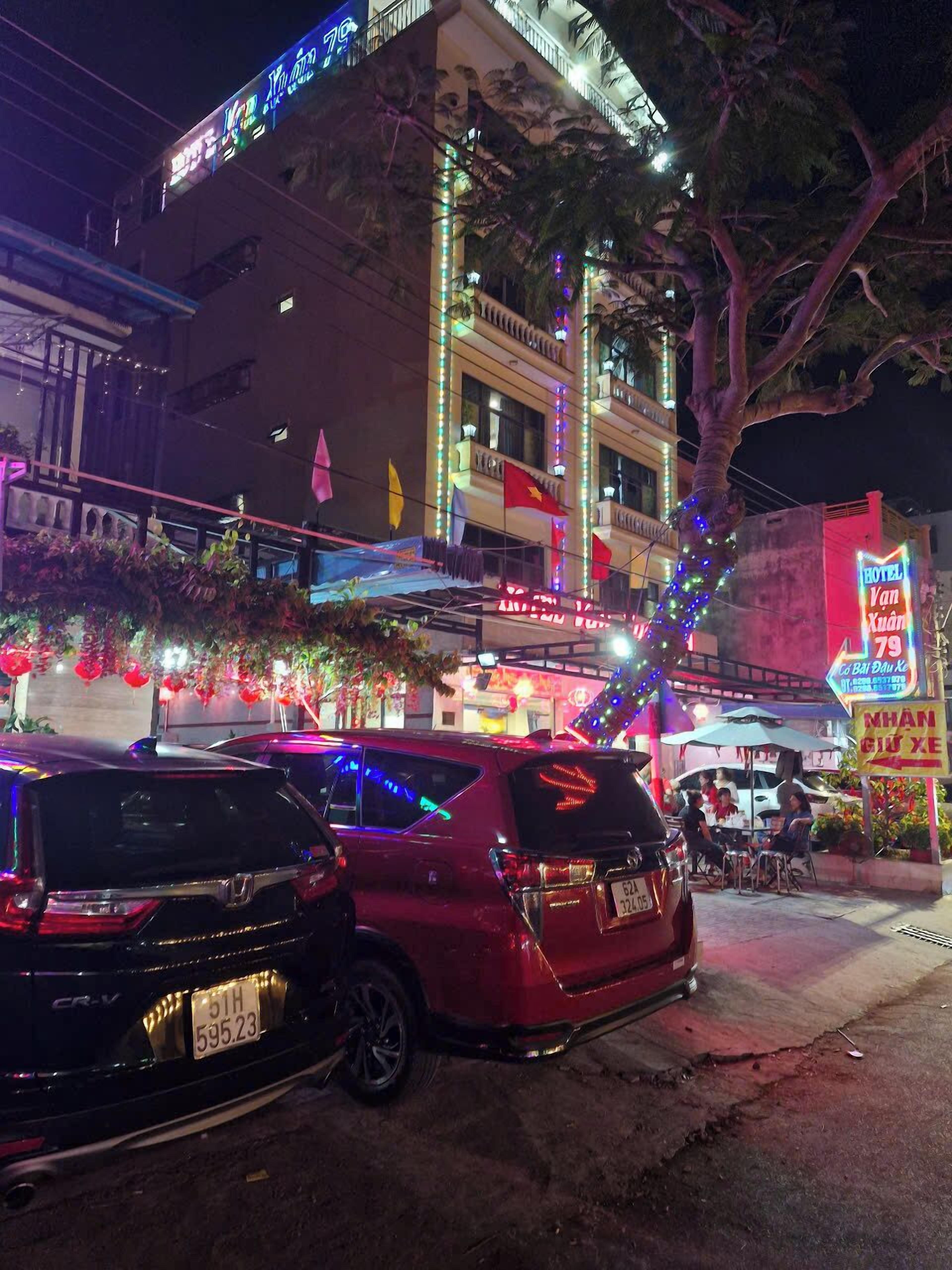 Un edificio con diversi piani, balconi e una segnaletica che indica che si tratta di un hotel. Diverse auto sono parcheggiate davanti e alcune persone siedono all'esterno.