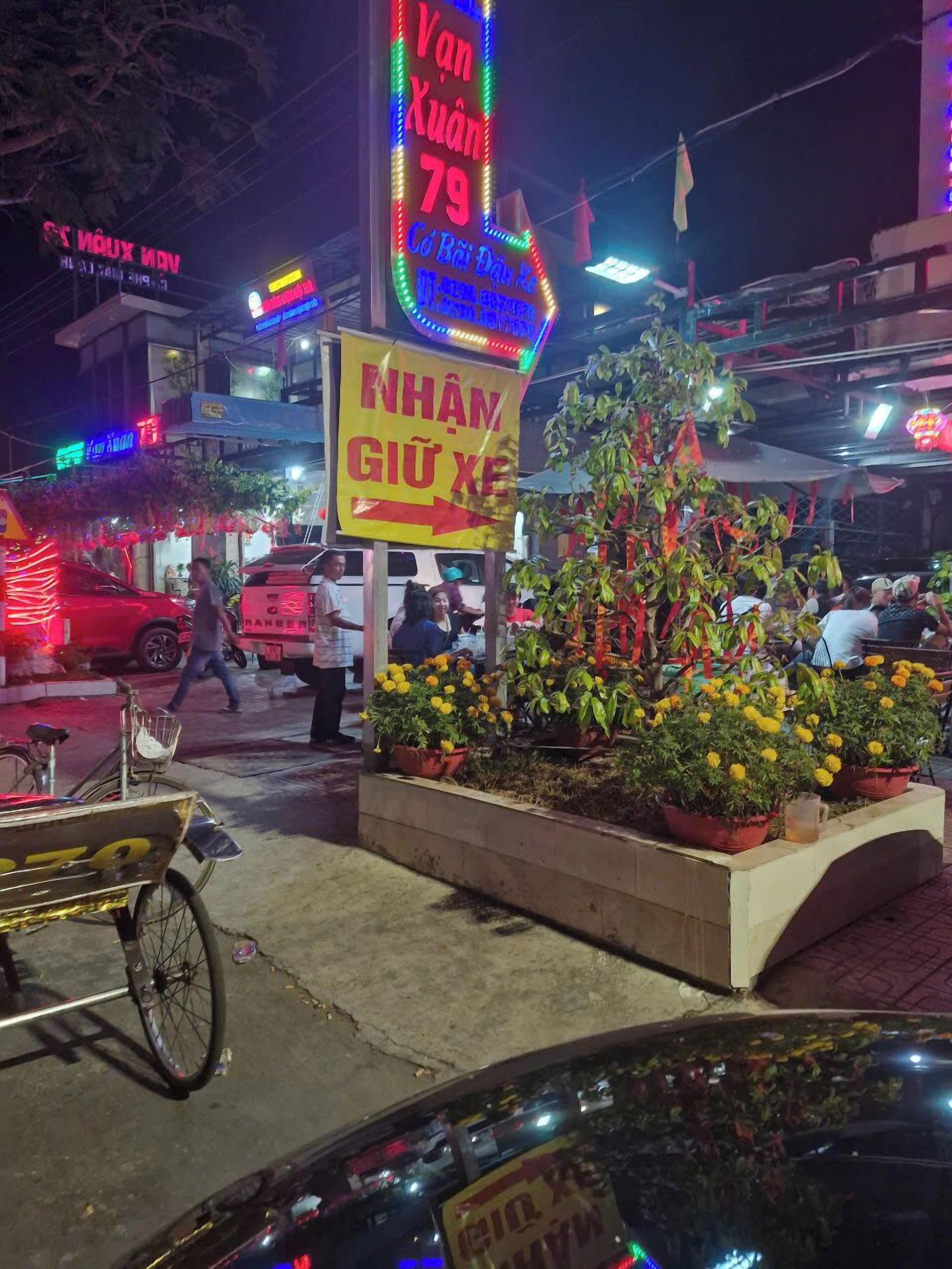 A street scene with a neon sign for a restaurant, a bicycle, and potted plants.
