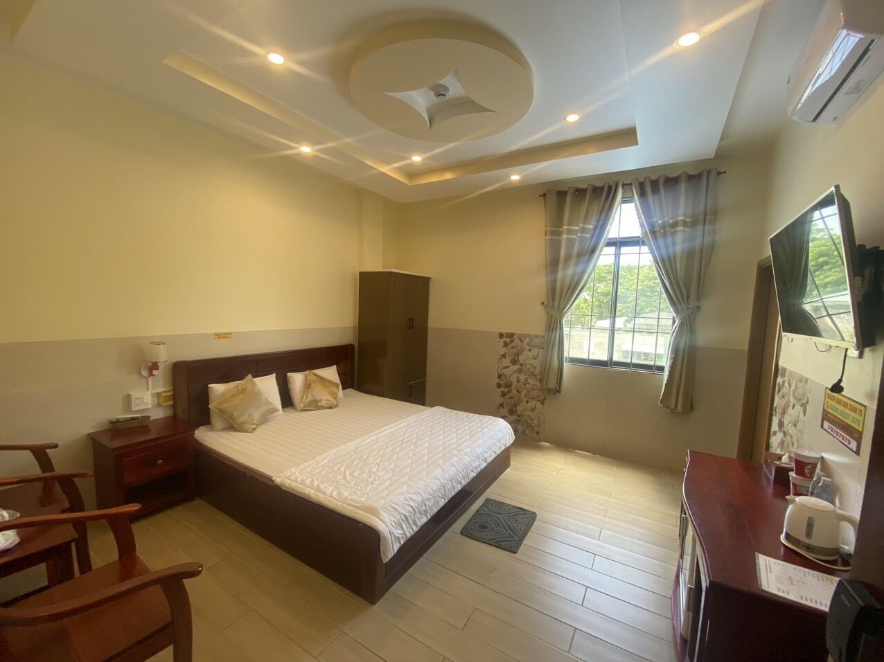 A hotel room with a large bed, a desk with a chair, a wardrobe, a window with curtains, and a ceiling fan.