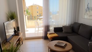 Una sala de estar con sofá, mesa de centro, televisor y balcón con vista a edificios y una palmera.