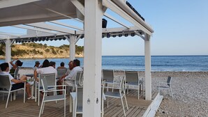 Un espacio de estar al aire libre cubierto con sillas y mesas de metal blanco, con vistas a una playa de costa rocosa y al mar.