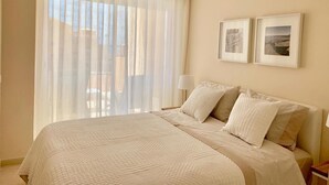Un dormitorio con una cama grande, cortinas translúcidas, dos cuadros en la pared y una mesita de noche con lámpara.