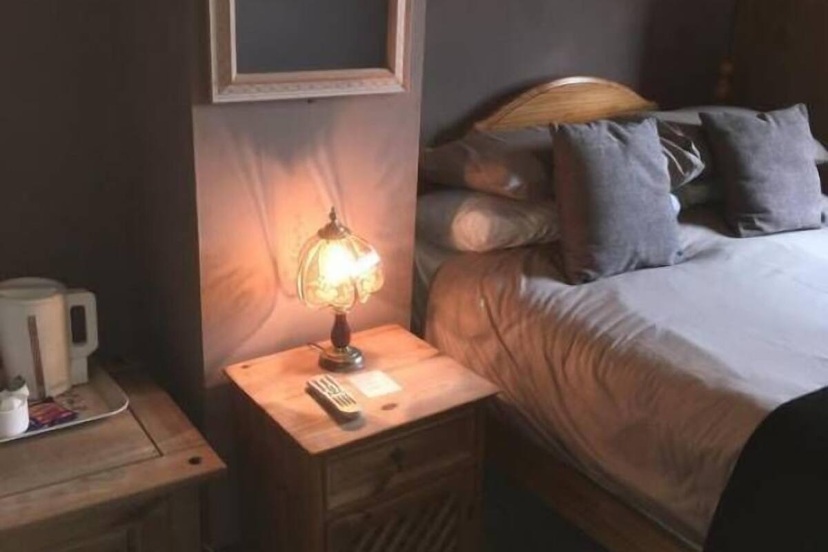 Un lit bien fait, avec une literie grise et une tête de lit en bois. On aperçoit une table de chevet avec une lampe et une télécommande.