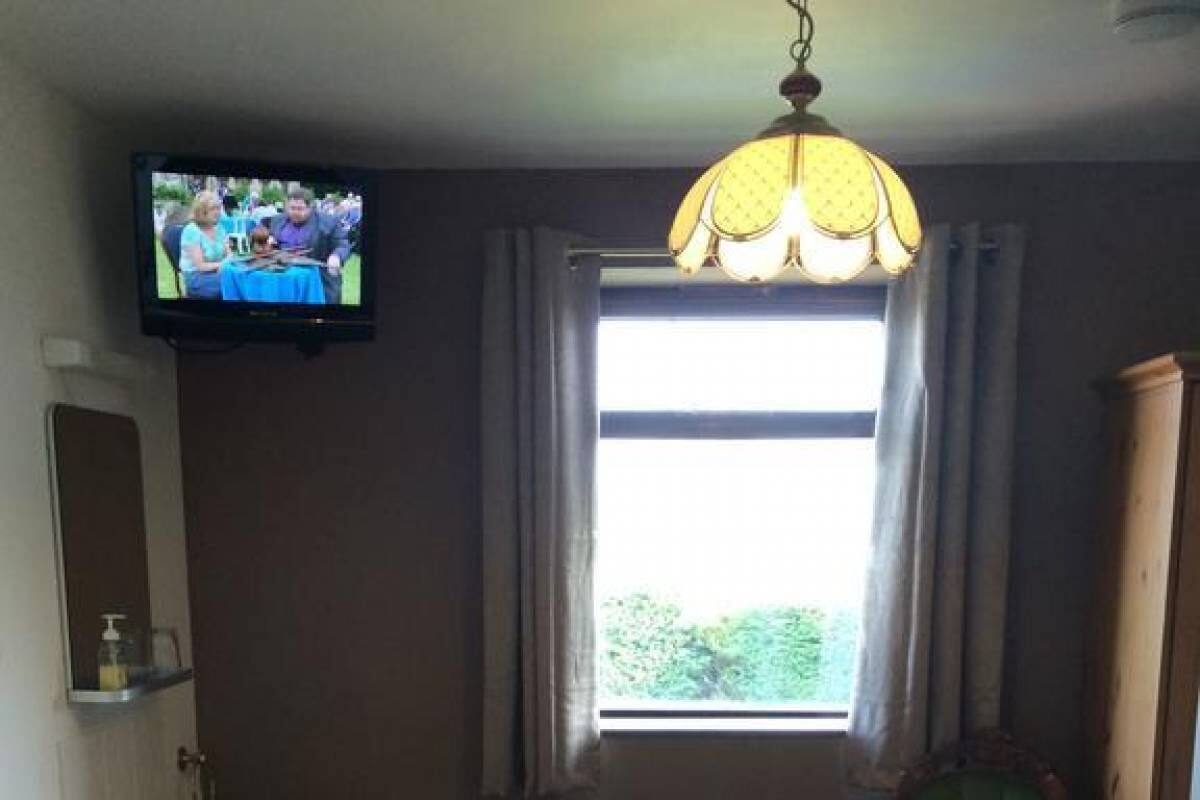 Une chambre à coucher avec un lit, une commode en bois, un fauteuil vert, une télévision, une fenêtre avec des rideaux et un radiateur.